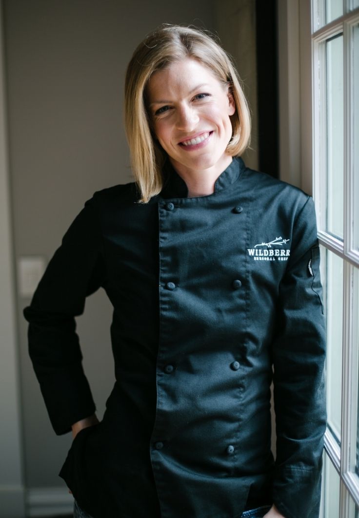 a woman standing next to a window wearing a black chef's coat with white writing on it