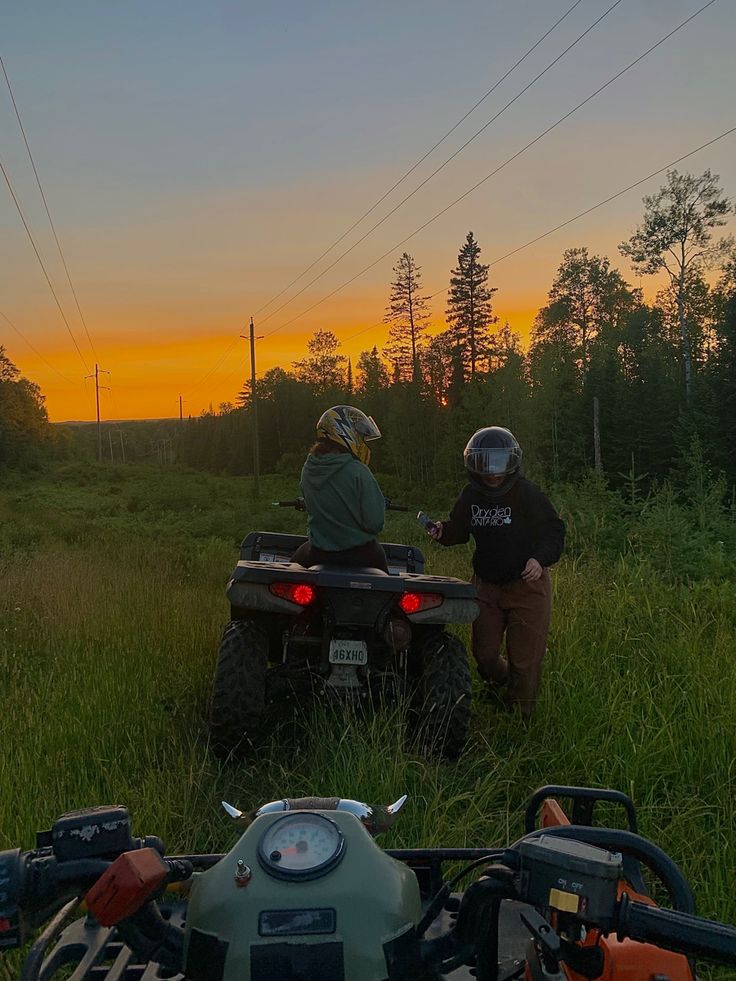 two people riding on four wheelers in the grass at sunset with trees and power lines behind them