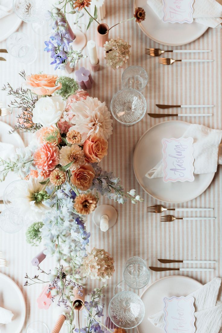 the table is set with white plates and flowers on it, along with place settings