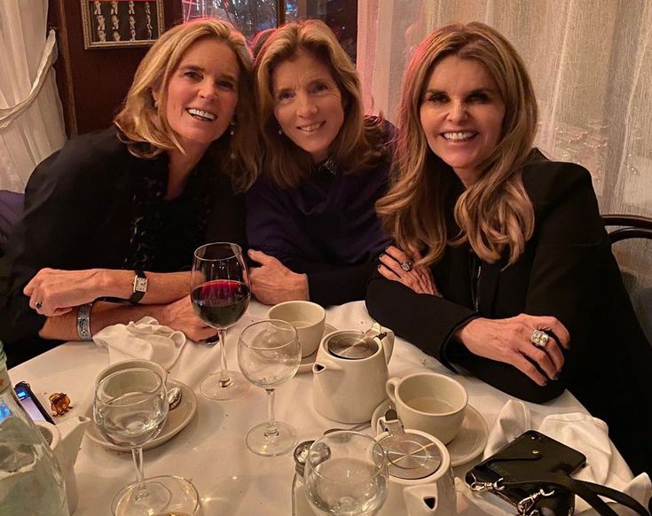 three women are sitting at a table with wine glasses and plates on it, posing for the camera