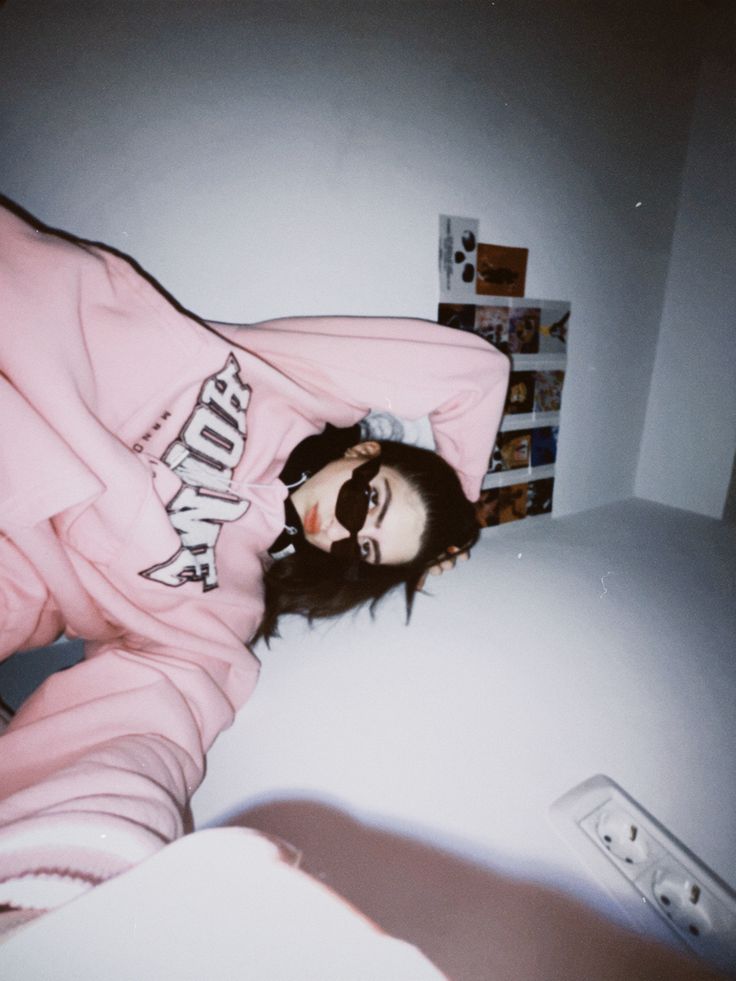 a woman laying on top of a bed wearing a pink sweatshirt and black sunglasses with her head resting on the pillow