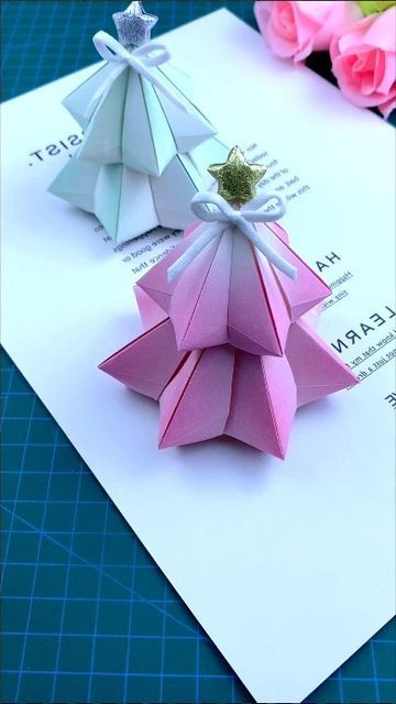 two origami christmas trees sitting on top of a piece of paper next to pink flowers