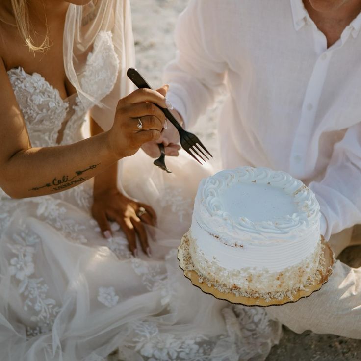 a man and woman cutting into a white cake on top of a sandy beach next to the ocean