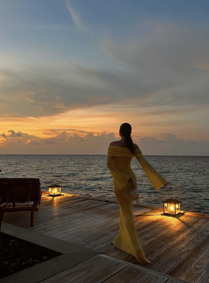 a woman in a long yellow dress standing on a dock at sunset with her arms outstretched