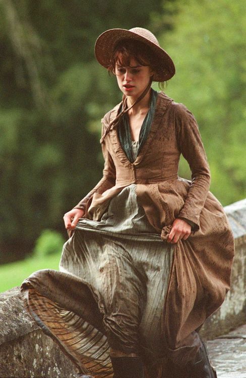 a woman in a brown dress and hat sitting on a rock