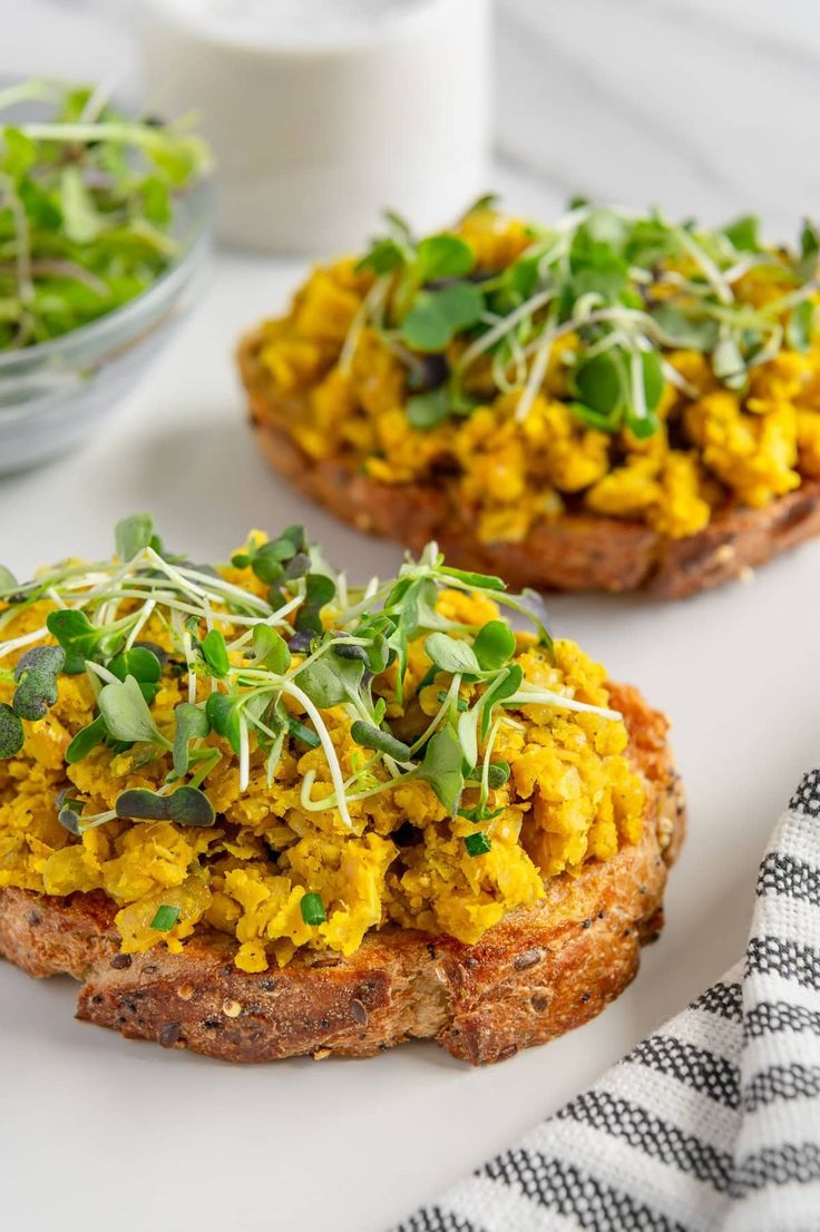 two pieces of bread topped with vegetables and sprouts next to a bowl of yogurt