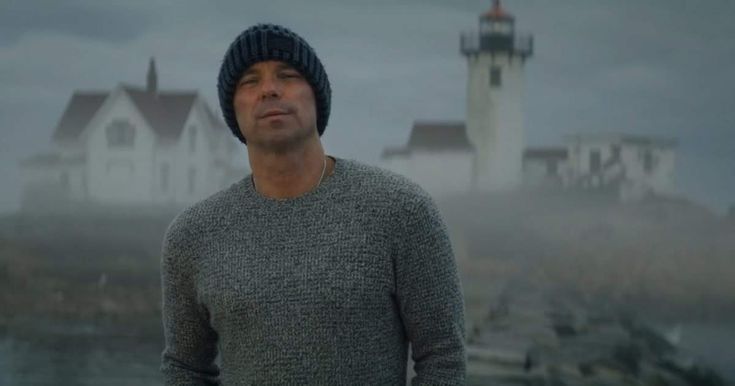 a man wearing a hat standing in front of a lighthouse on a foggy day