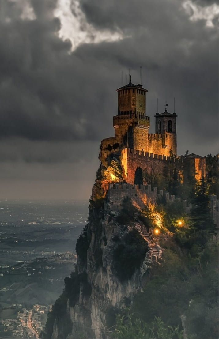 a castle on top of a mountain with dark clouds in the sky above it and lights below