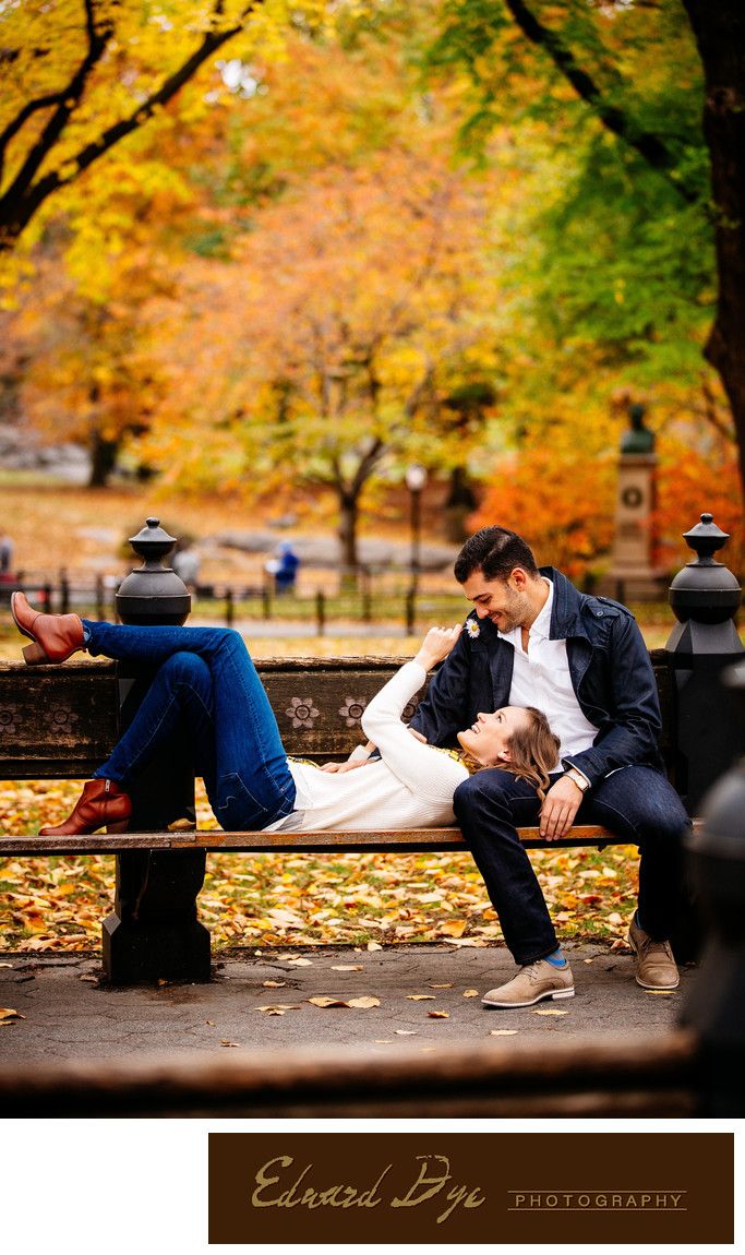 a man and woman sitting on a park bench in the fall with leaves all around them