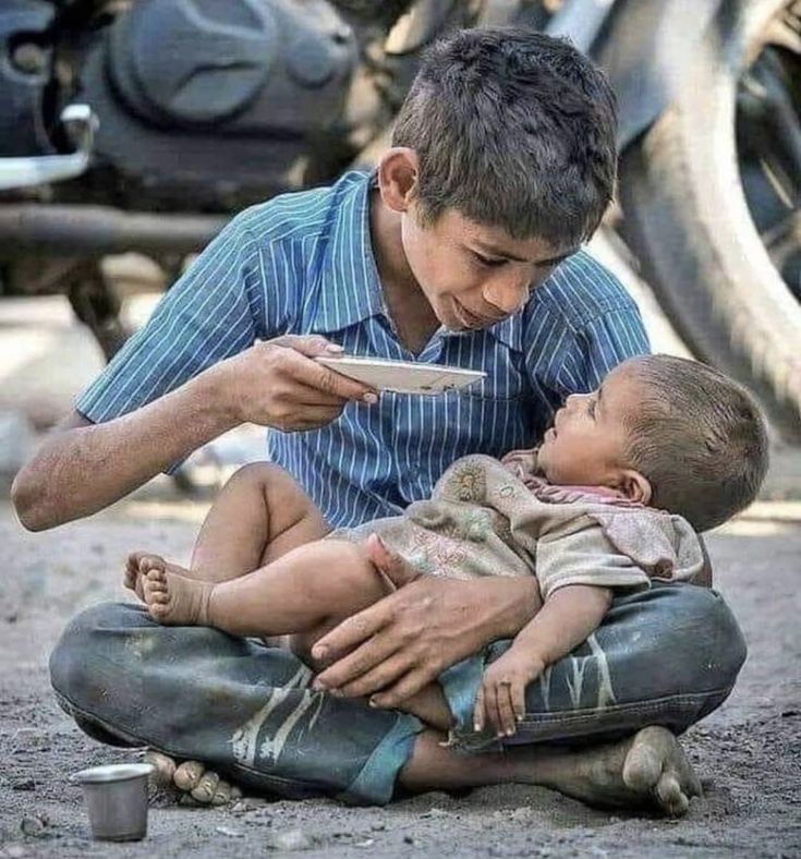 a man holding a baby and feeding it with a spoon while sitting on the ground