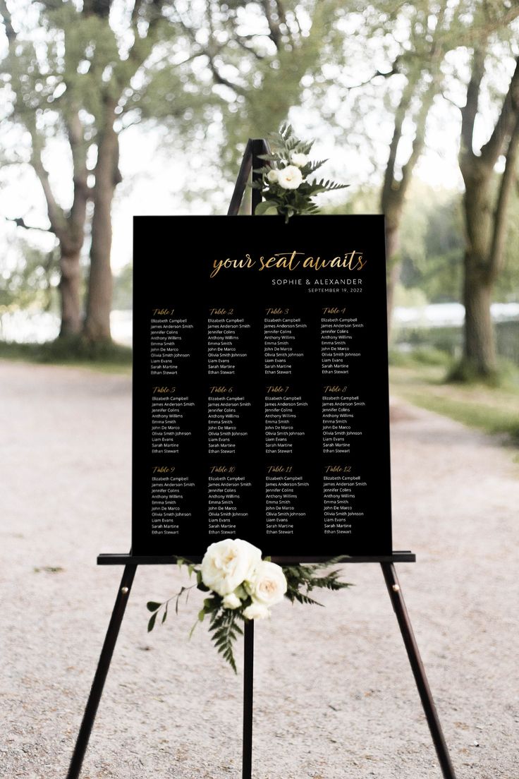 a black and white wedding seating sign with flowers on it in front of some trees