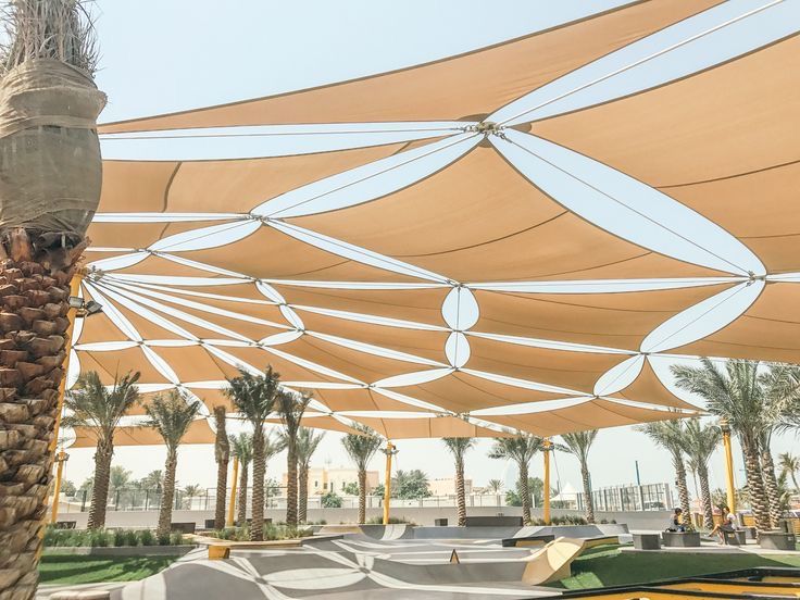 an outdoor covered area with benches and palm trees in the foreground, surrounded by large shade sails