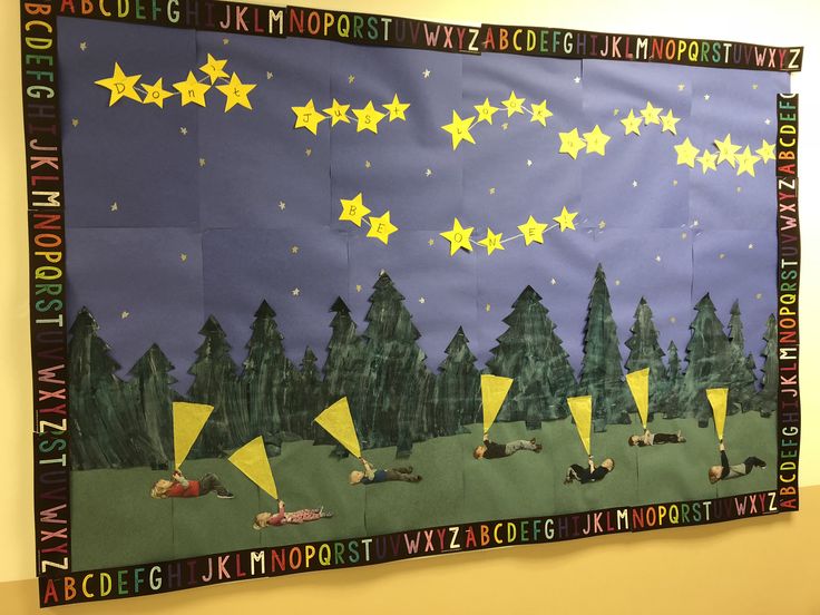 a bulletin board with yellow stars on it in the middle of a classroom wall that has trees and animals painted on it