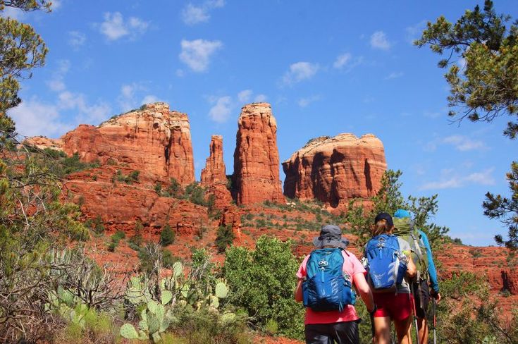 two people with backpacks are hiking in the desert near red rocks and cactus trees