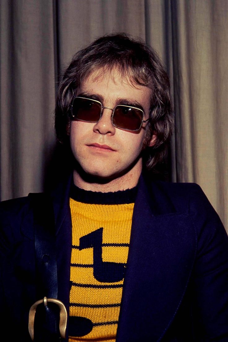 a man wearing sunglasses sitting in front of a curtain with his hands on his hips