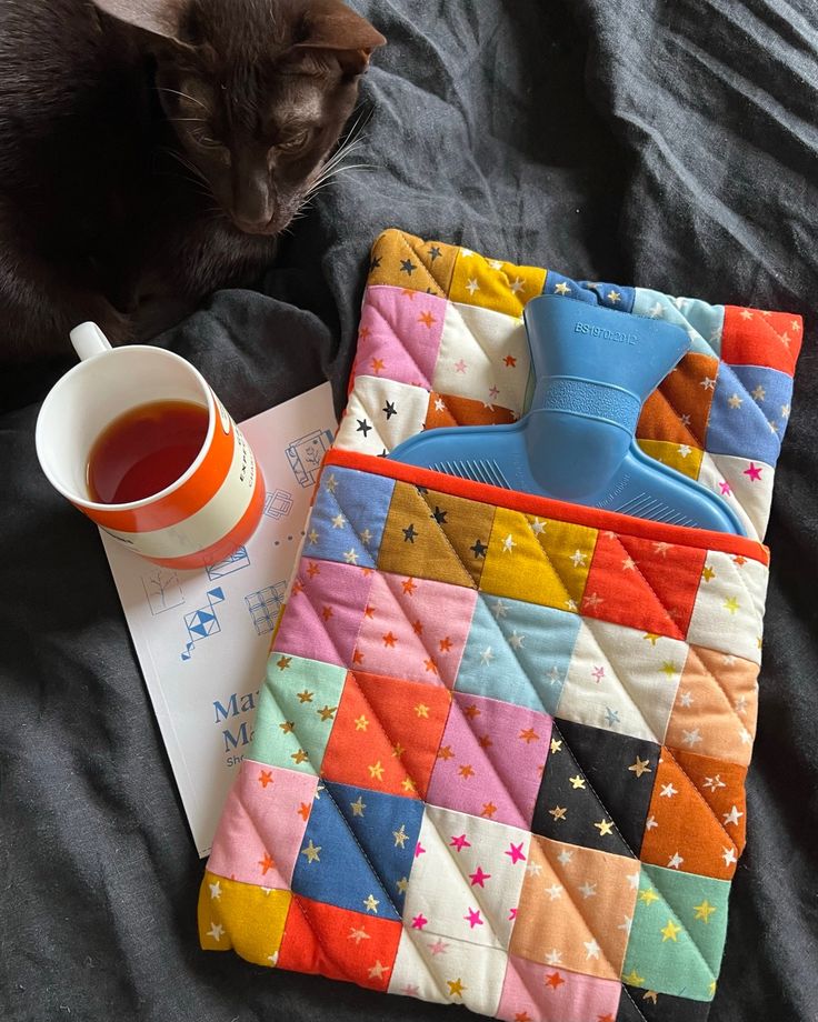 a cat laying on top of a bed next to a cup of tea and an oven mitt