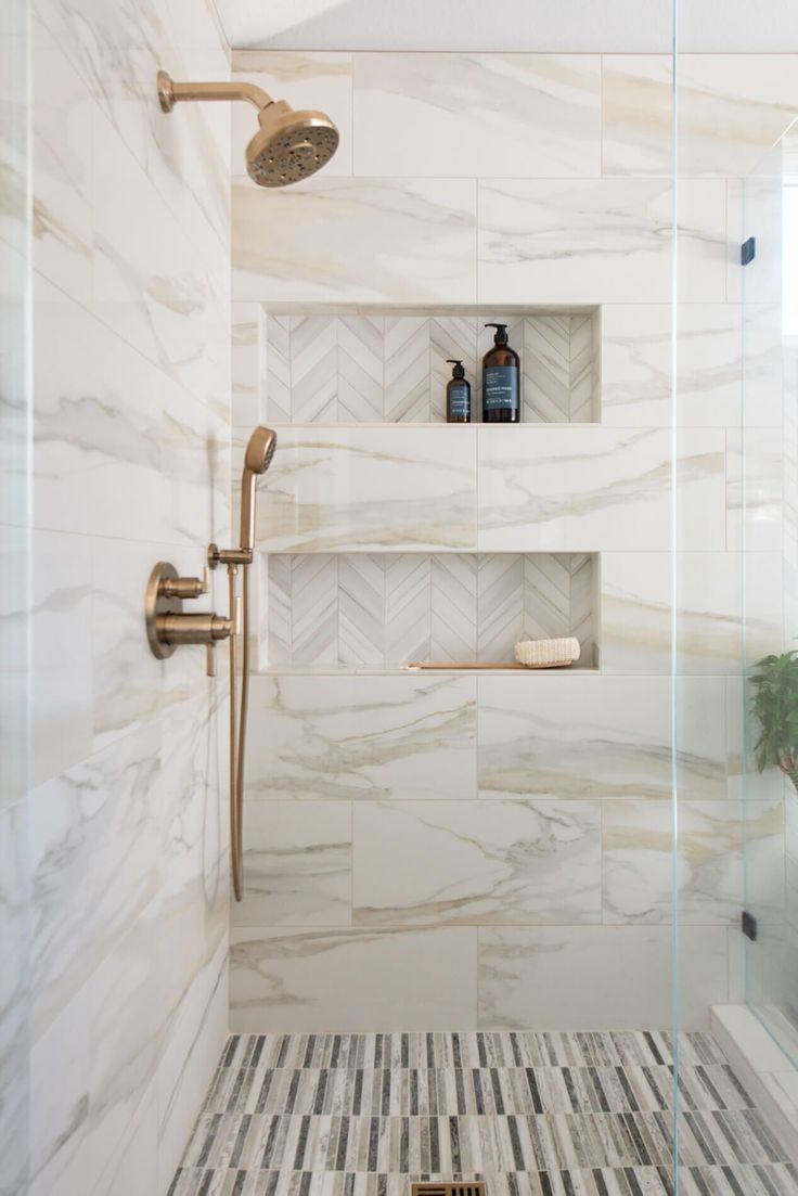 a bathroom with white marble and gold fixtures, shower head, toilet paper holder and shelves on the wall