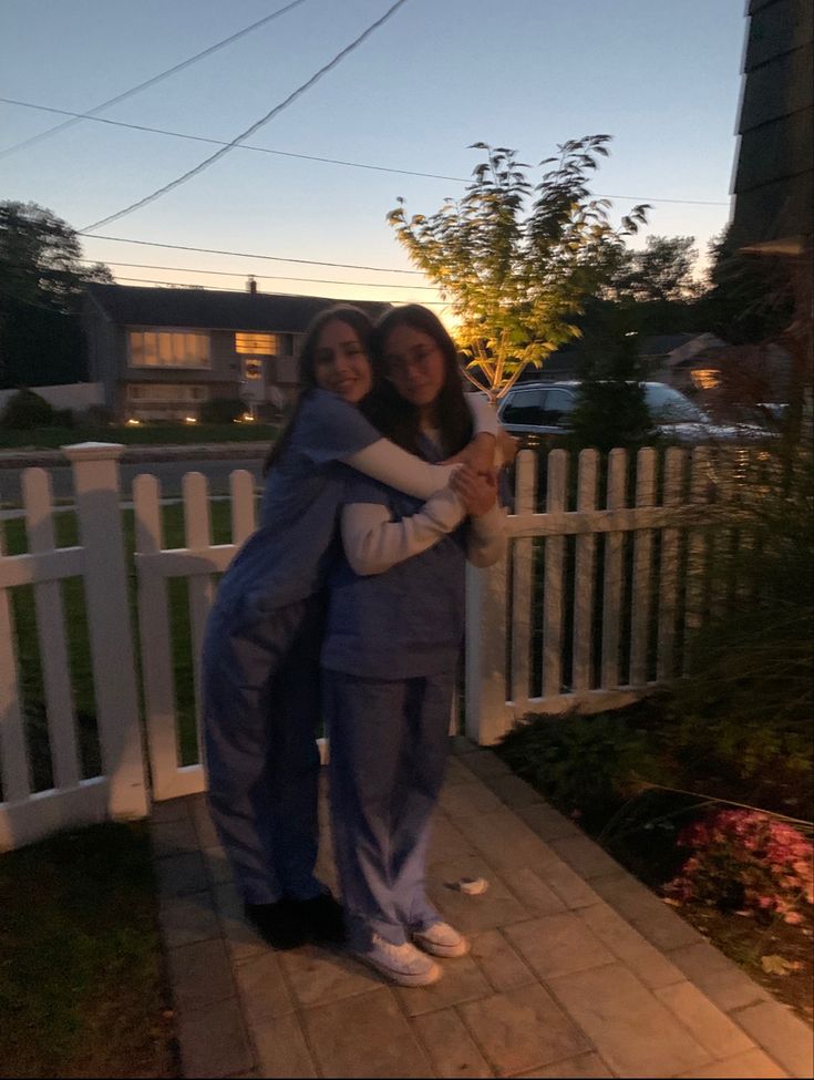 two women hugging each other in front of a white picket fence at night with the sun setting behind them