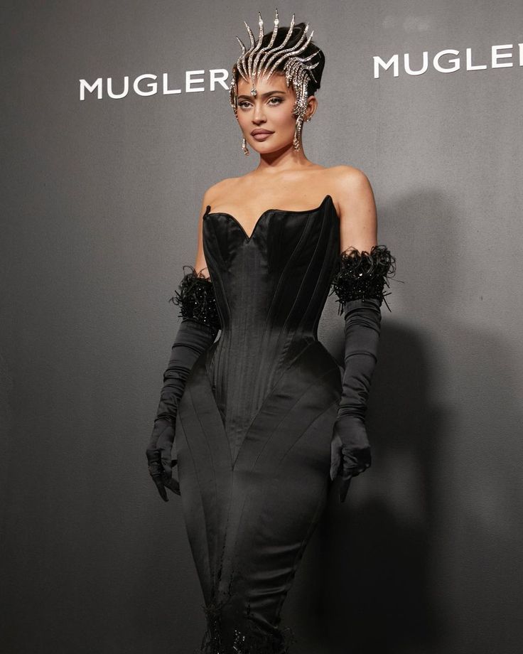 a woman in a black dress and gloves standing next to a sign that says mugler mugler