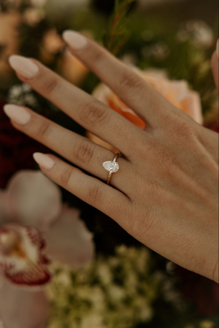 a woman's hand with an engagement ring on top of her finger and flowers in the background