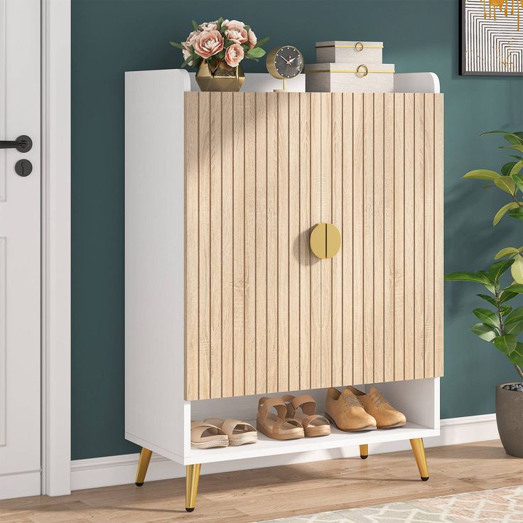 a white cabinet with wooden doors and shoes on the bottom shelf in front of a potted plant