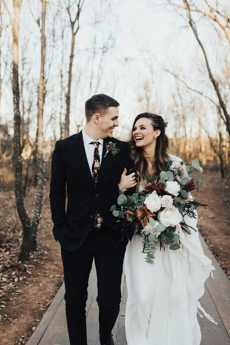 a bride and groom are walking down the path in front of some trees with their bouquets