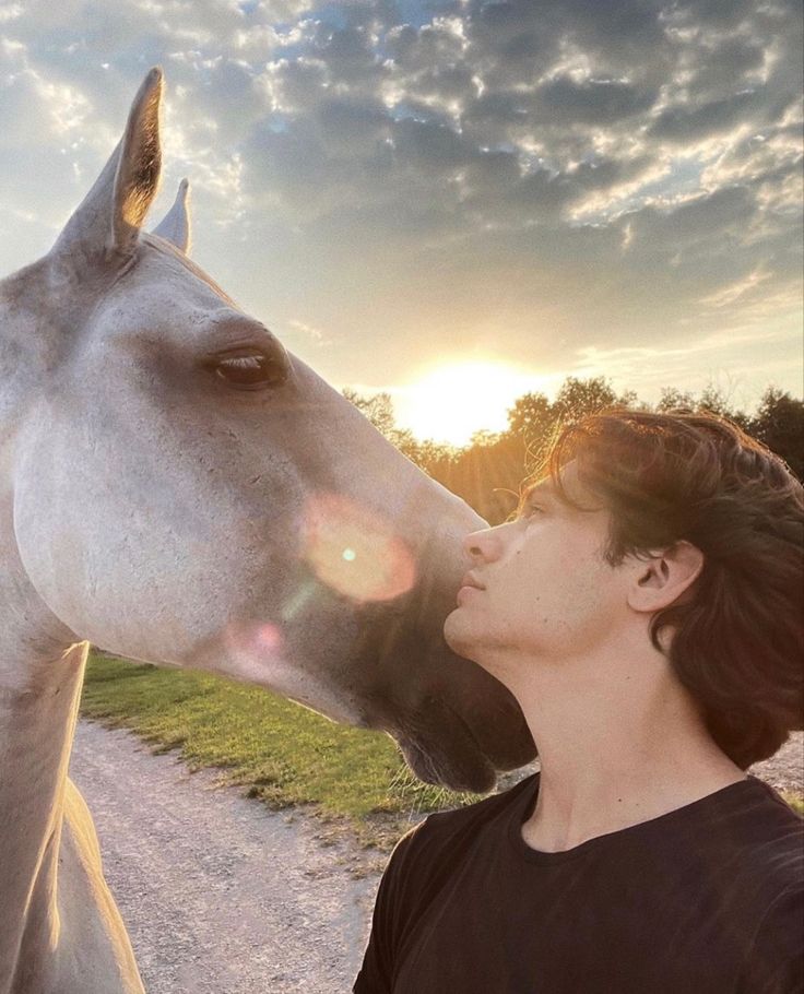 a woman standing next to a white horse on a dirt road with the sun behind her