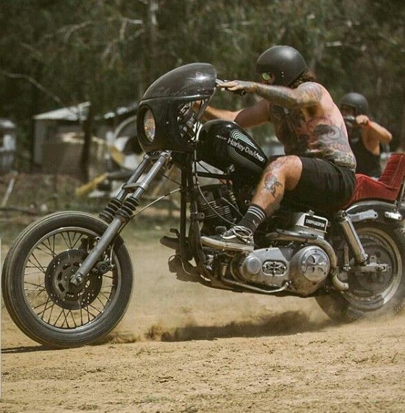 a tattooed man riding on the back of a motorcycle down a dirt road next to trees