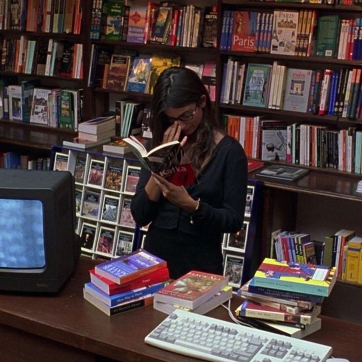 a woman sitting at a desk reading a book in front of a computer and bookshelf