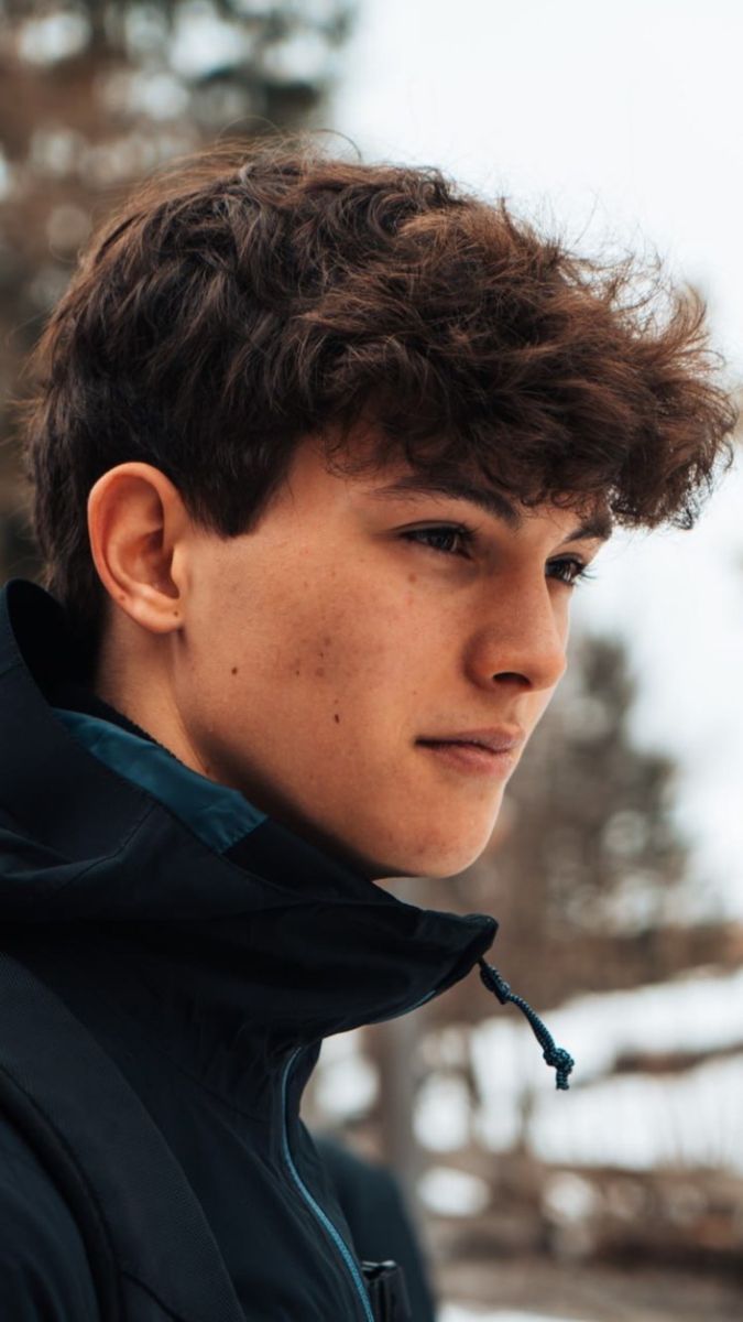 a young man with freckles on his hair looks at the camera while standing in front of snow covered trees