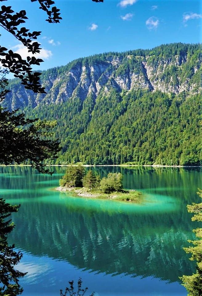 a lake surrounded by mountains and trees with green water in the foreground, under a blue sky