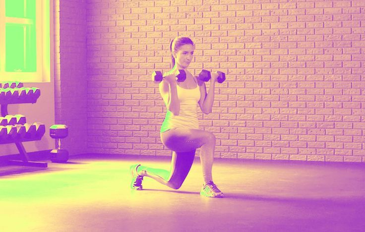 a woman squatting with two dumbbells in front of a brick wall and exercise equipment behind her