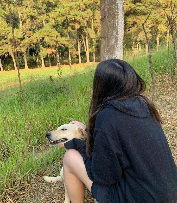 a woman sitting on the ground with her dog in front of some trees and grass