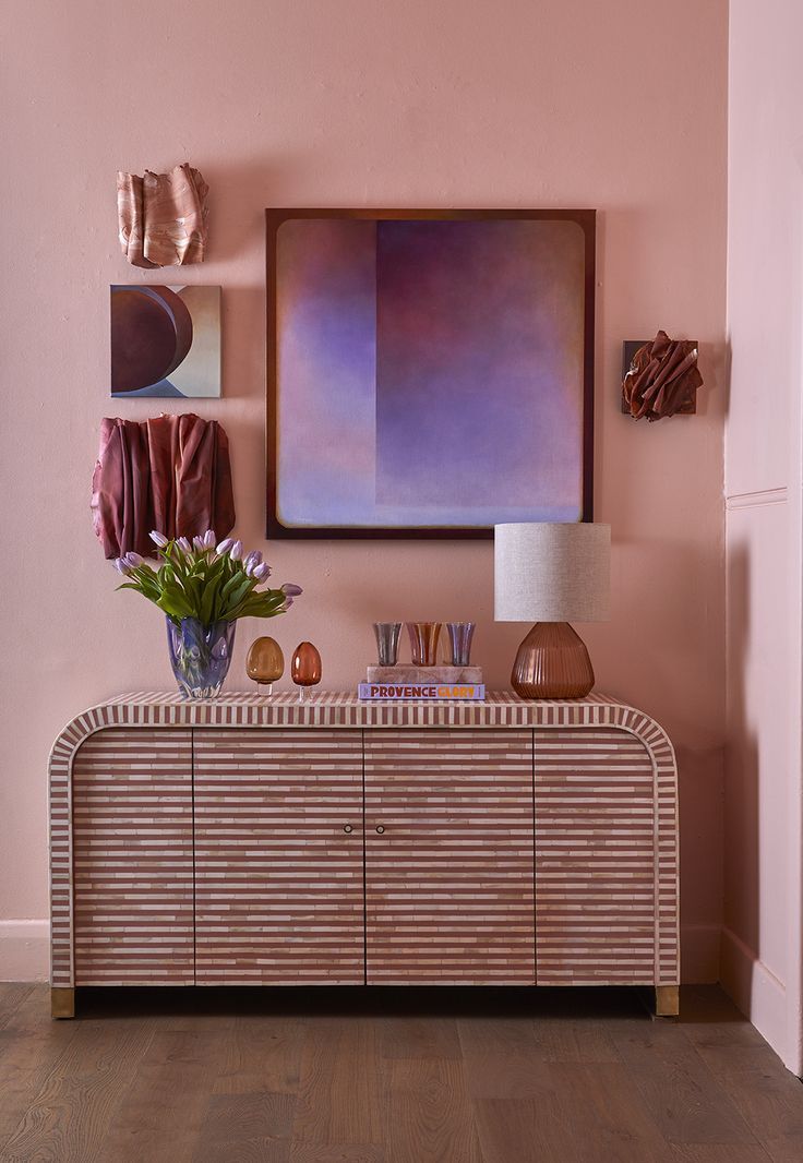 a room with pink walls and an art piece on the wall above a buffet cabinet