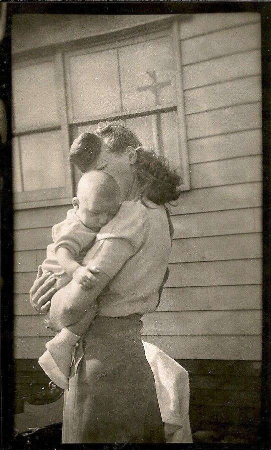 an old black and white photo of a woman holding a baby in front of a house