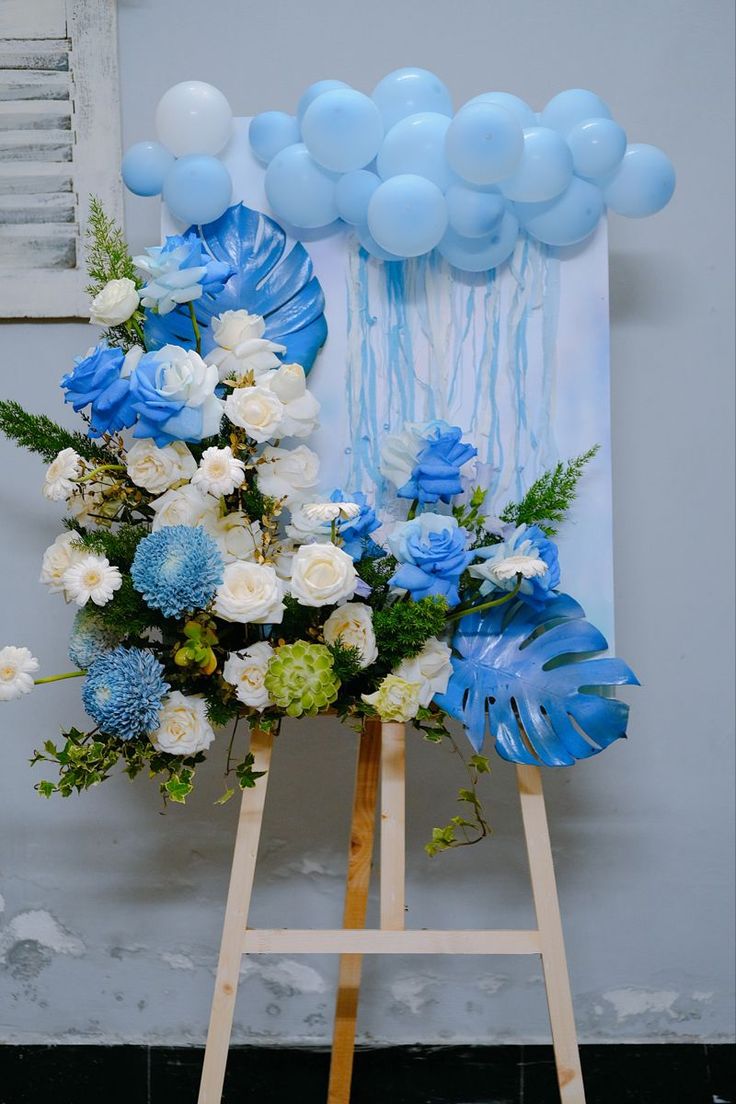 a blue and white floral arrangement on an easel with balloons in the air behind it