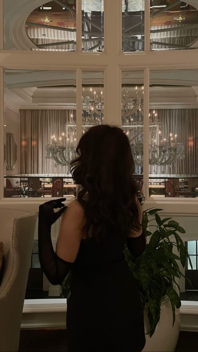 a woman standing in front of a large window looking out at the outside world with chandeliers on either side