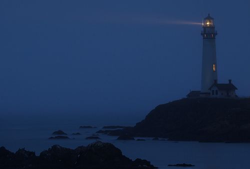 the light house is lit up at night on the rocky shore by the water's edge