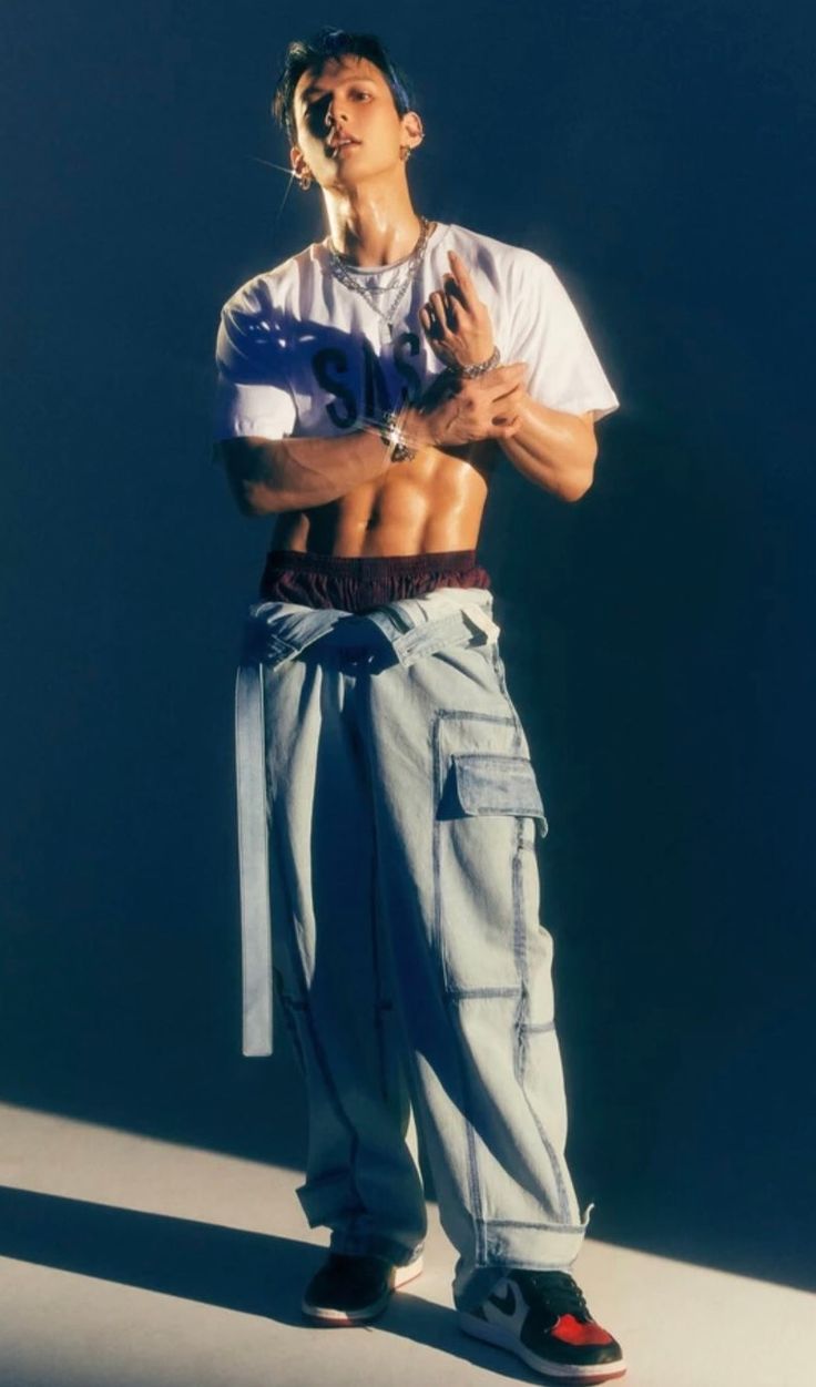 a man with no shirt on standing in front of a wall wearing blue cargo pants