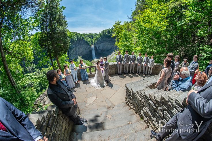 a group of people standing around each other in front of some trees and rocks with a waterfall behind them