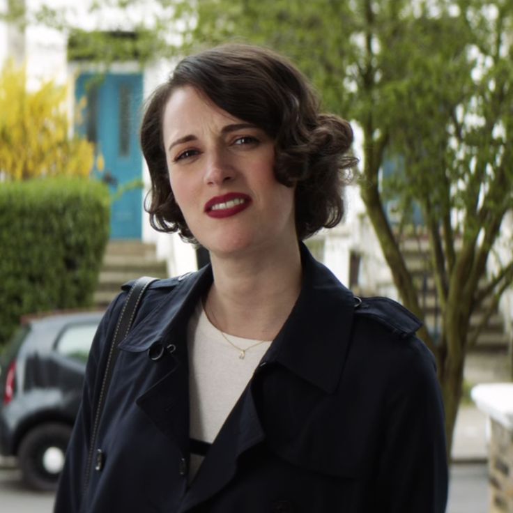 a woman standing on the street in front of a building with cars and trees behind her