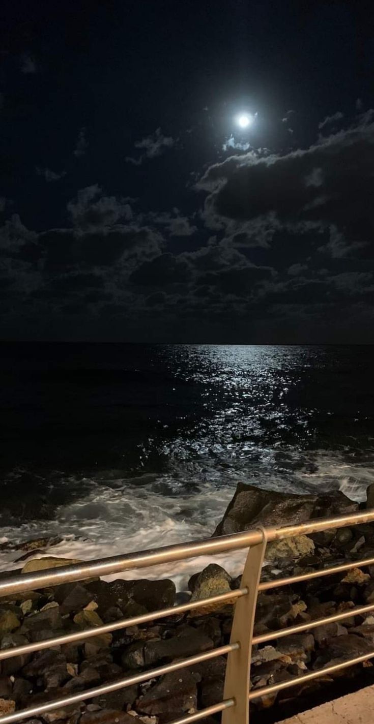 a full moon is seen over the ocean and rocky shore at night with clouds in the sky