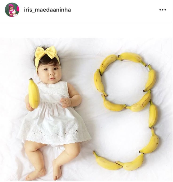 a baby laying next to two bananas on a white sheet and wearing a yellow bow