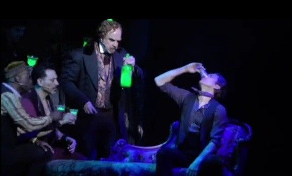 two men in suits and one is holding a green bottle while another man sits on a couch