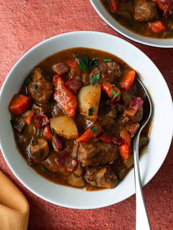 two white bowls filled with stew on top of a red tablecloth next to a spoon