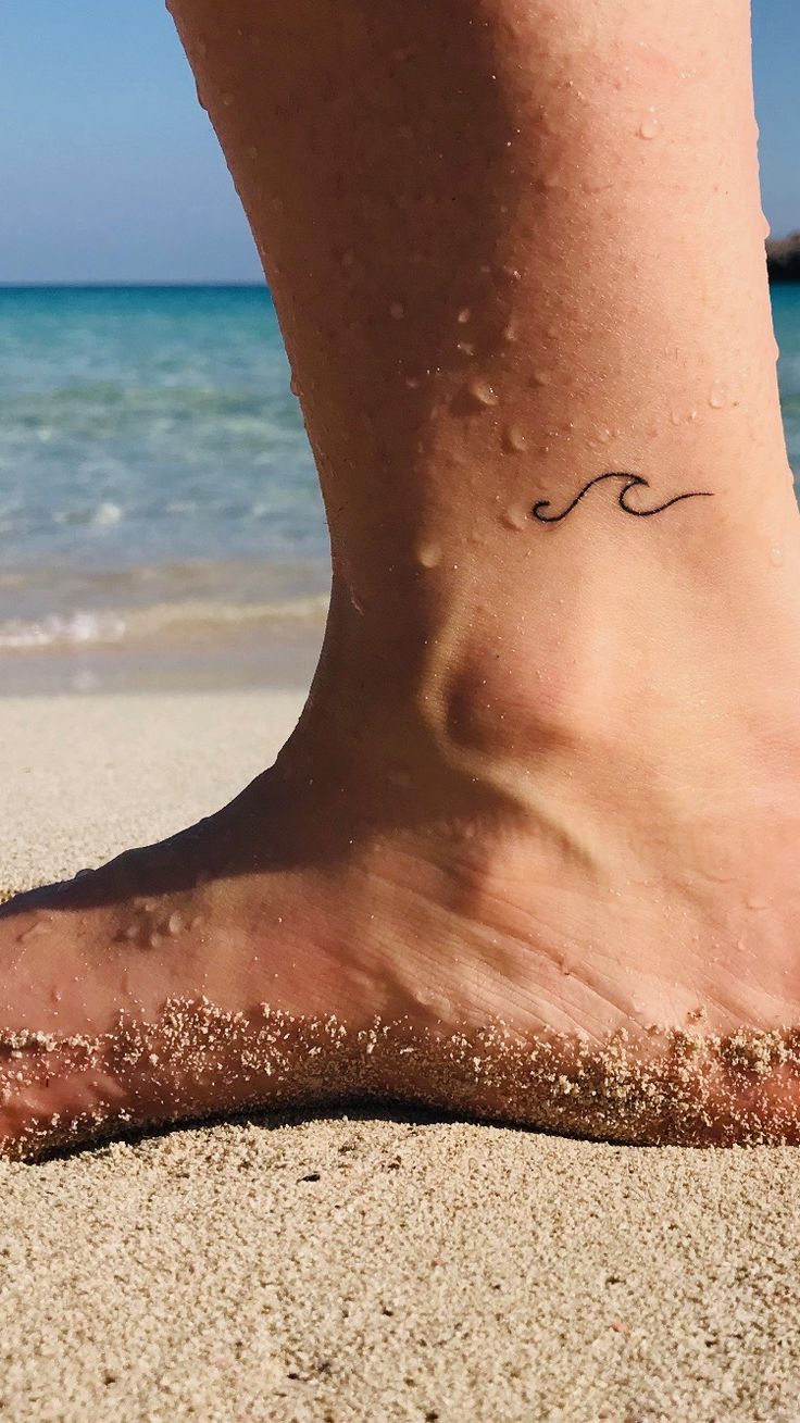 a person's foot with a wave tattoo on the sand at the beach by the ocean