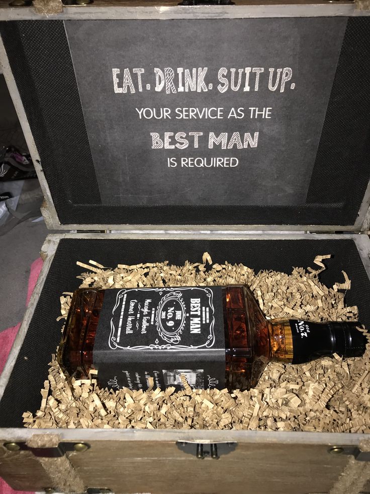 an empty beer bottle sitting in a wooden box filled with wood shavings next to a sign that says eat drink suit up, your service as the best man is required