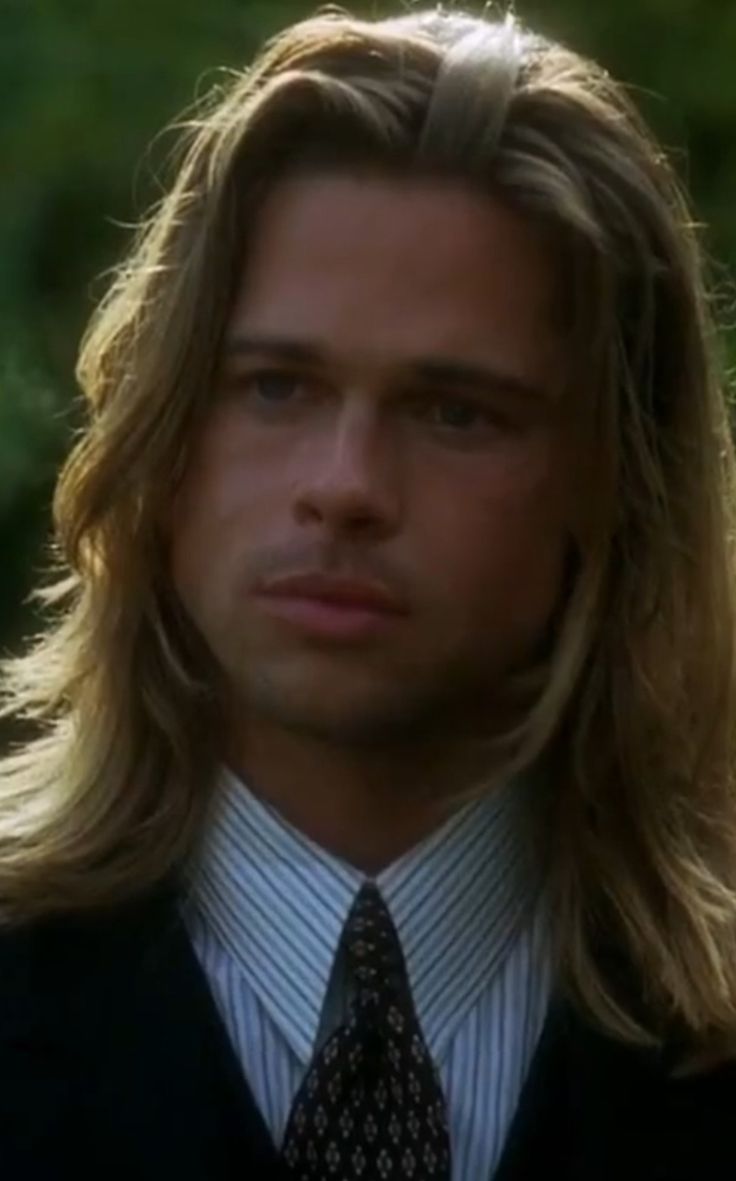 a man with long hair wearing a suit and tie looking at the camera while standing in front of trees