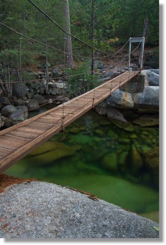 a wooden bridge over a river surrounded by rocks and trees with green water in the foreground