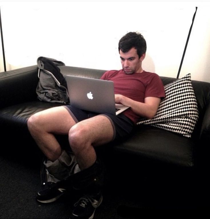 a man sitting on a couch using a laptop computer with his legs crossed and holding a backpack